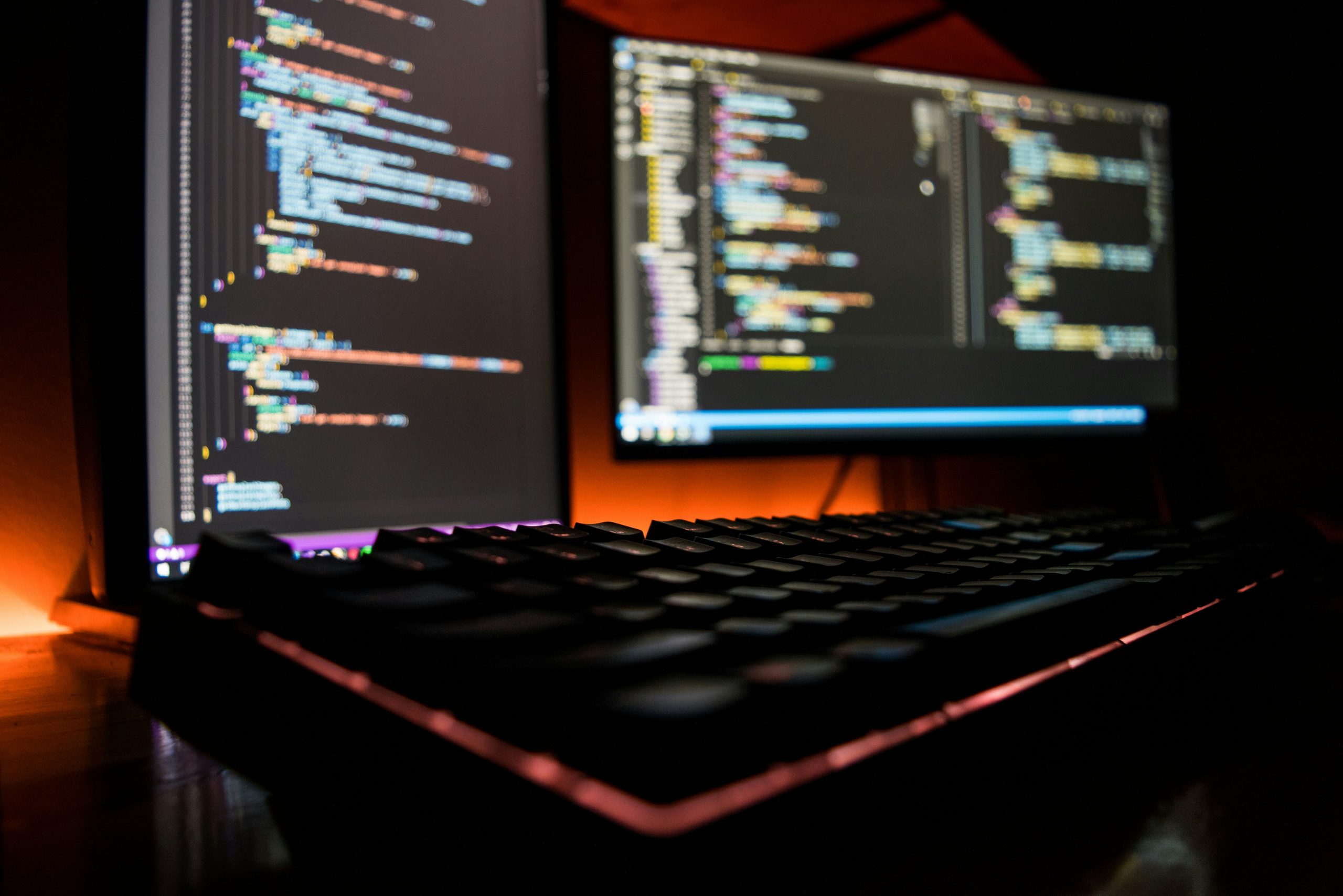 close-up shot of a mechanical keyboard with RGB lighting in the foreground, with two blurred computer monitors displaying lines of colored code in a dark room in the background.